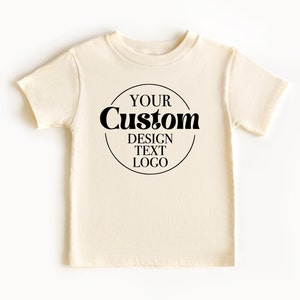 Op de afbeelding: Een cr&egrave;mekleurig T-shirt met een zwarte cirkelvormige afbeelding met de tekst "YOUR Custom DESIGN TEXT LOGO".