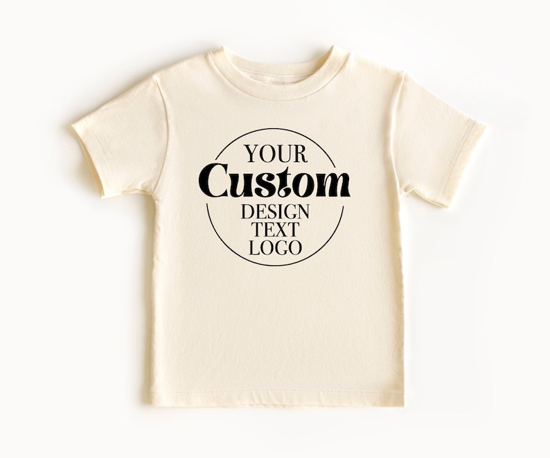 Op de afbeelding: Een cr&egrave;mekleurig T-shirt met een zwarte cirkelvormige afbeelding met de tekst "YOUR Custom DESIGN TEXT LOGO".