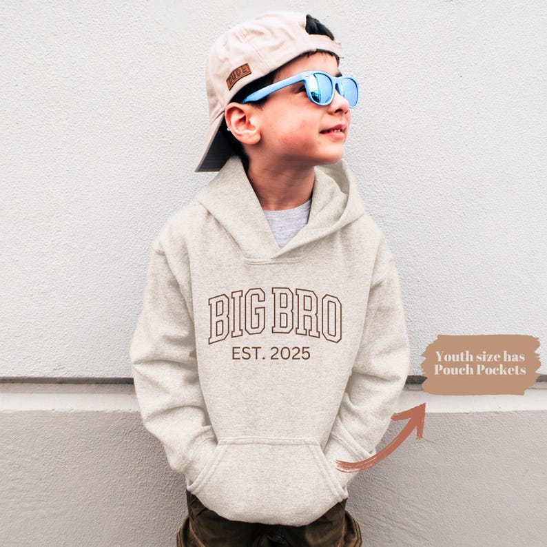 K&ouml;nnte beinhalten: Ein hellbeiger Hoodie mit einem braunen Umriss des Textes "BIG BRO EST. 2025" auf der Vorderseite. Ein junger Mensch tr&auml;gt den Hoodie und eine hellbraune Baseballkappe mit blauer Sonnenbrille.