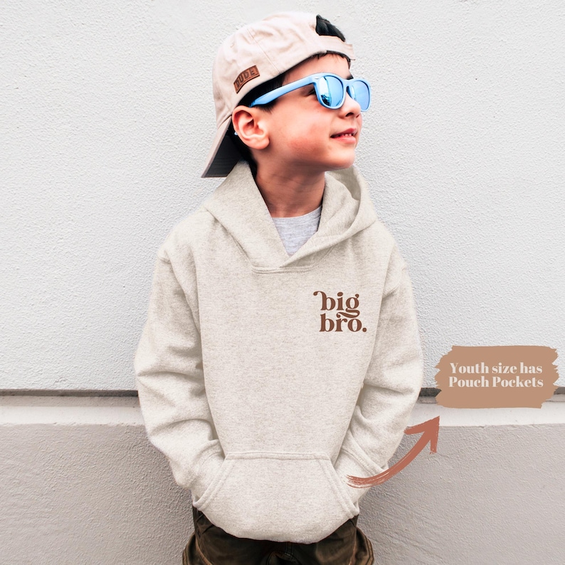 May include: A light beige hoodie with a brown 'big bro.' graphic on the front. The hoodie has a pouch pocket and is being worn by a child wearing a light blue baseball cap and blue sunglasses.