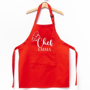 K&ouml;nnte beinhalten: Rote K&uuml;chensch&uuml;rze mit einer wei&szlig;en Kochm&uuml;tze und dem Text "Chef Emma" auf der Vorderseite.