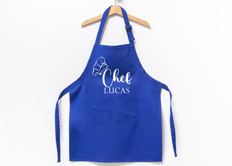K&ouml;nnte beinhalten: Blaue Kochsch&uuml;rze mit einer wei&szlig;en Kochm&uuml;tze und dem Text "Chef Lucas" auf der Vorderseite.