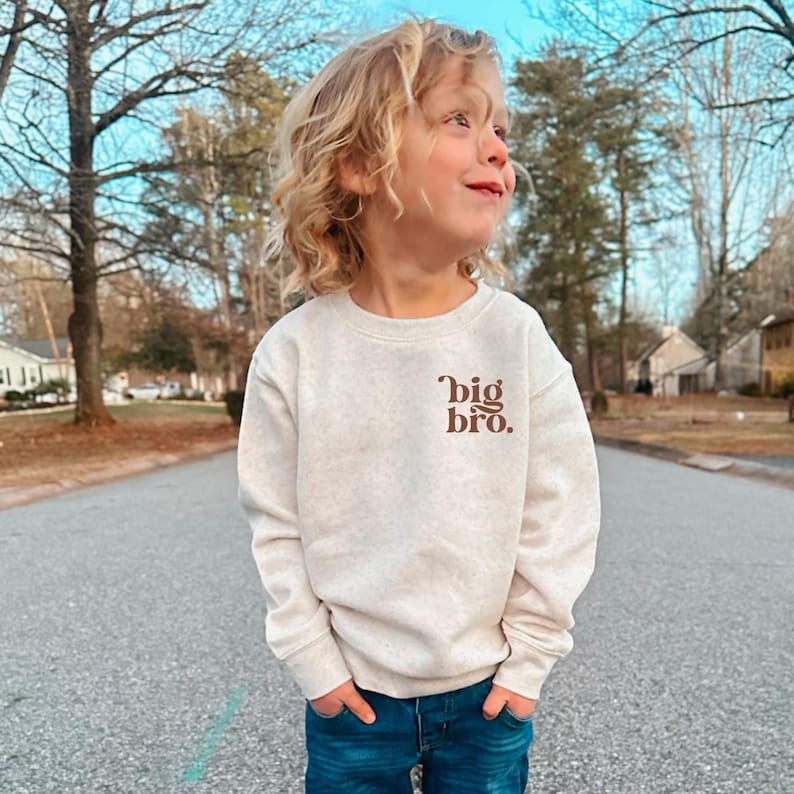 May include: A young child wearing a light beige sweatshirt with the text "big bro." printed in brown on the front. The child is standing on a paved road with trees in the background.