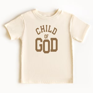 May include: A cream colored short-sleeved t-shirt with the words "CHILD OF GOD" printed in brown on the front.