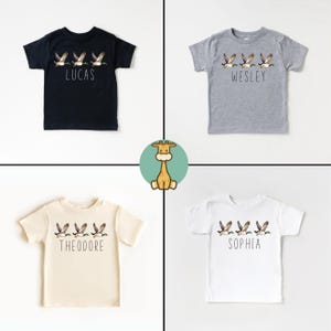 May include: Four white t-shirts with a line of flying ducks and a name printed below. The shirts are in black, grey, cream, and white.