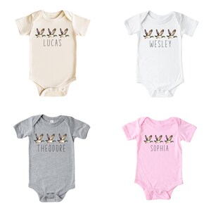 May include: Four baby onesies with a line of flying ducks and a name printed below. The onesies are in beige, white, grey, and pink.
