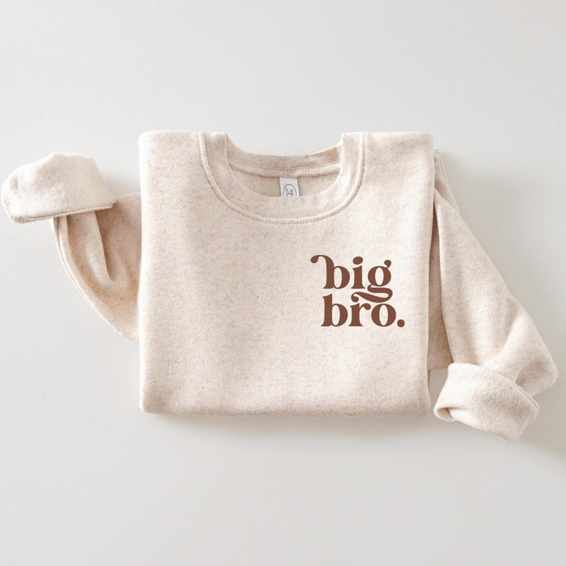 Puede incluir: Una sudadera beige con un gr&aacute;fico marr&oacute;n que dice "big bro."