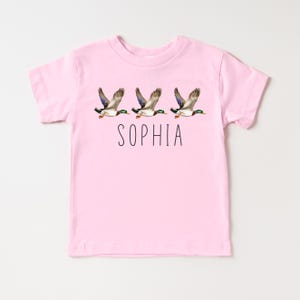 May include: Pink t-shirt with a graphic of three flying ducks and the name "SOPHIA" printed below.