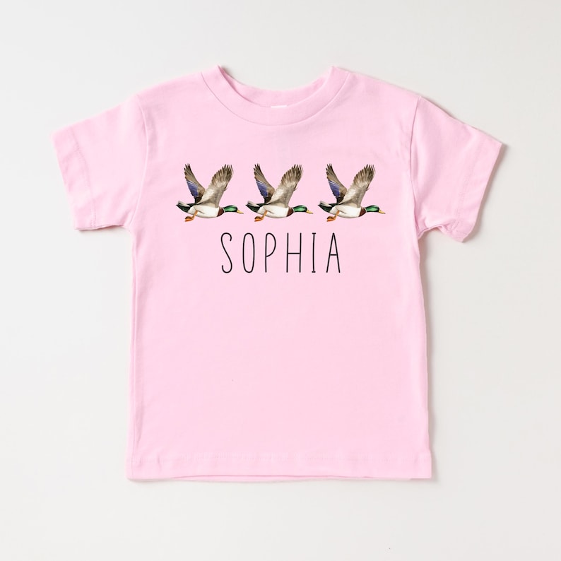May include: Pink t-shirt with a graphic of three flying ducks and the name "SOPHIA" printed below.