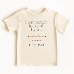 以下が含まれることがあります： クリーム色のTシャツに、「hand picked for earth by my custom name in heaven」という黒い文字がプリントされています。 「custom」という単語は赤色で、両側に小さな赤いハートが付いています。