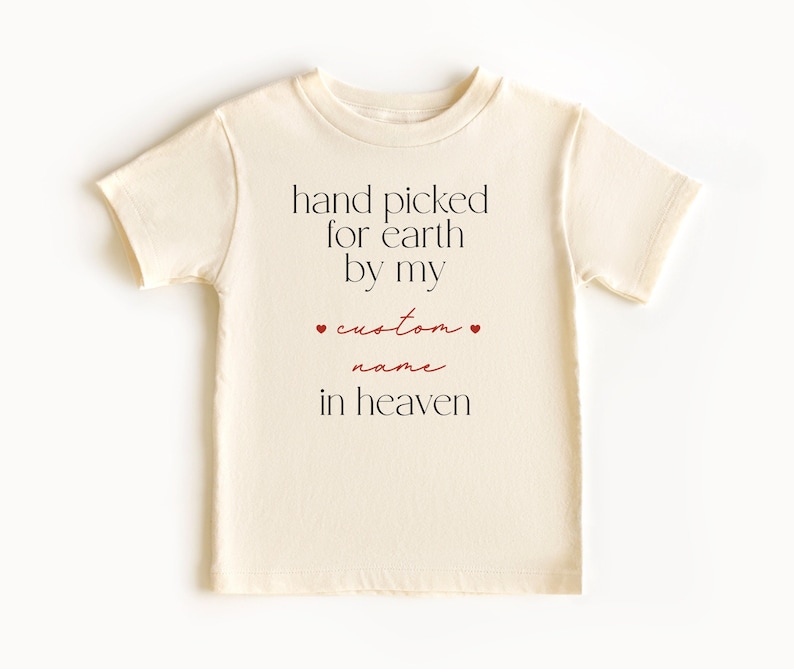 以下が含まれることがあります： クリーム色のTシャツに、「hand picked for earth by my custom name in heaven」という黒い文字がプリントされています。 「custom」という単語は赤色で、両側に小さな赤いハートが付いています。