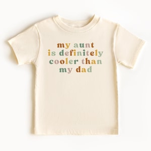 May include: A cream colored t-shirt with the text "my aunt is definitely cooler than my dad" printed in colorful letters.