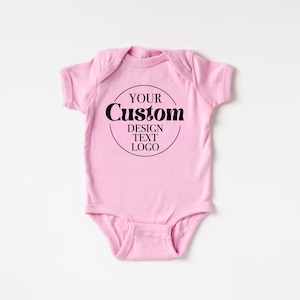 Op de afbeelding: Roze baby onesie met een zwarte cirkel en de tekst "YOUR CUSTOM DESIGN TEXT LOGO" in een retro lettertype.