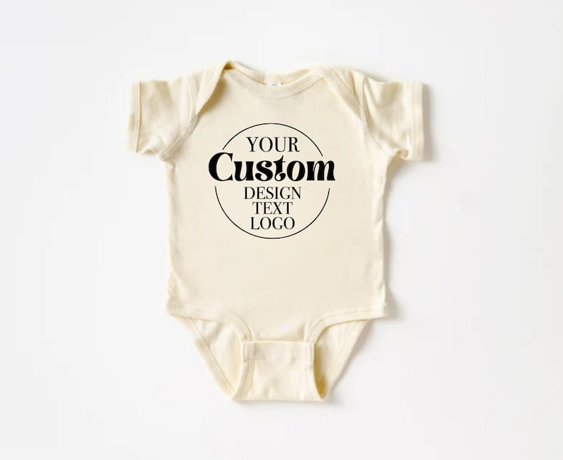 Op de afbeelding: Een cr&egrave;mekleurige baby romper met een zwarte cirkel met de tekst "YOUR Custom DESIGN TEXT LOGO".