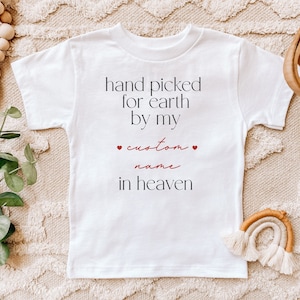 以下が含まれることがあります： 白いベビーロンパースに「hand picked for earth by my custom name in heaven」という黒い文字と赤いハートが2つ書かれています。