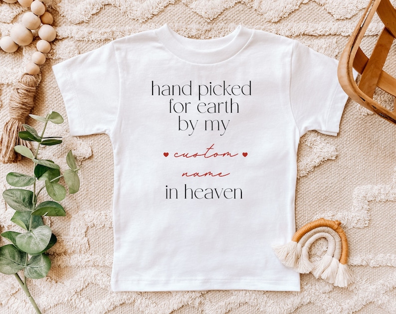 以下が含まれることがあります： 白いベビーロンパースに「hand picked for earth by my custom name in heaven」という黒い文字と赤いハートが2つ書かれています。