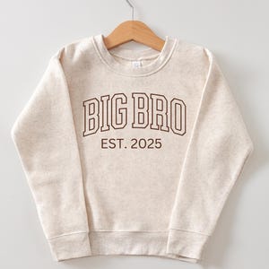 K&ouml;nnte beinhalten: Ein beiger Pullover mit dem Text "BIG BRO EST. 2025" in Braun.