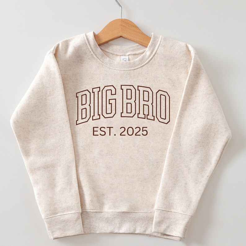 K&ouml;nnte beinhalten: Ein beiger Pullover mit dem Text "BIG BRO EST. 2025" in Braun.