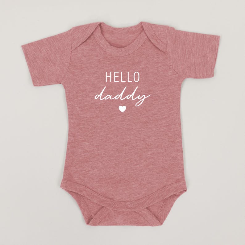May include: A pink baby onesie with short sleeves and the text "HELLO daddy" in white with a small white heart.