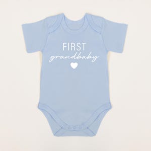 May include: A light blue baby onesie with the text "FIRST grandbaby" in white with a white heart.