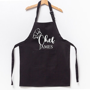 K&ouml;nnte beinhalten: Schwarze Sch&uuml;rze mit einer wei&szlig;en Kochm&uuml;tze und dem aufgedruckten Text "Chef James".