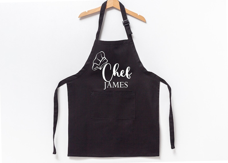 K&ouml;nnte beinhalten: Schwarze Sch&uuml;rze mit einer wei&szlig;en Kochm&uuml;tze und dem aufgedruckten Text "Chef James".