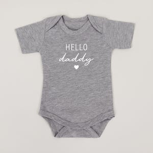 May include: Gray short-sleeved baby onesie with white text that reads "HELLO daddy" and a small white heart.