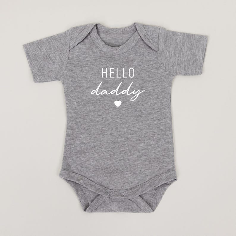 May include: Gray short-sleeved baby onesie with white text that reads "HELLO daddy" and a small white heart.