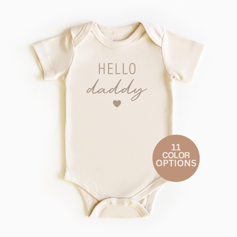 May include: A cream colored baby onesie with the text "HELLO daddy" in brown lettering and a small heart.