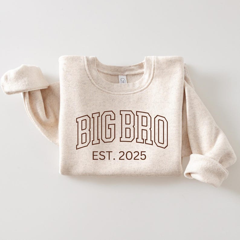 K&ouml;nnte beinhalten: Ein beiger Pullover mit dem Text "BIG BRO EST. 2025" in Braun.