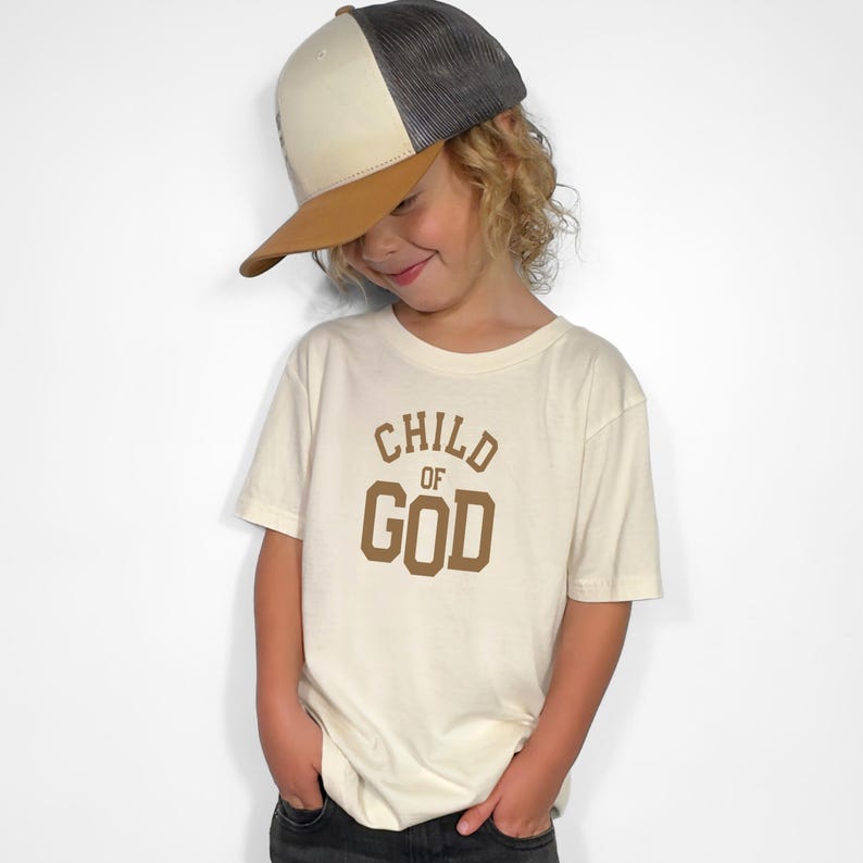 May include: A child wearing a white t-shirt with the text "CHILD OF GOD" in brown lettering. The child is also wearing a brown and white trucker hat.