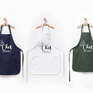 K&ouml;nnte beinhalten: Drei personalisierte Kochsch&uuml;rzen in Marineblau, Wei&szlig; und Gr&uuml;n mit dem Text "Chef" und einem Namen darunter.