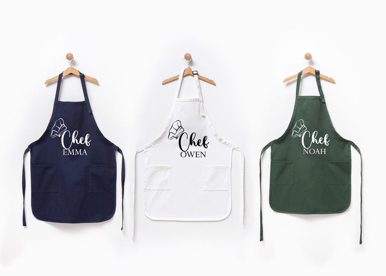 K&ouml;nnte beinhalten: Drei personalisierte Kochsch&uuml;rzen in Marineblau, Wei&szlig; und Gr&uuml;n mit dem Text "Chef" und einem Namen darunter.