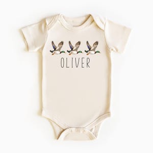 May include: A cream-colored baby onesie with a graphic of three ducks flying in a row. The onesie has the name "OLIVER" printed below the ducks.