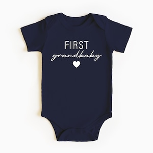 May include: Navy blue baby onesie with white text that reads "FIRST grandbaby" and a small white heart.