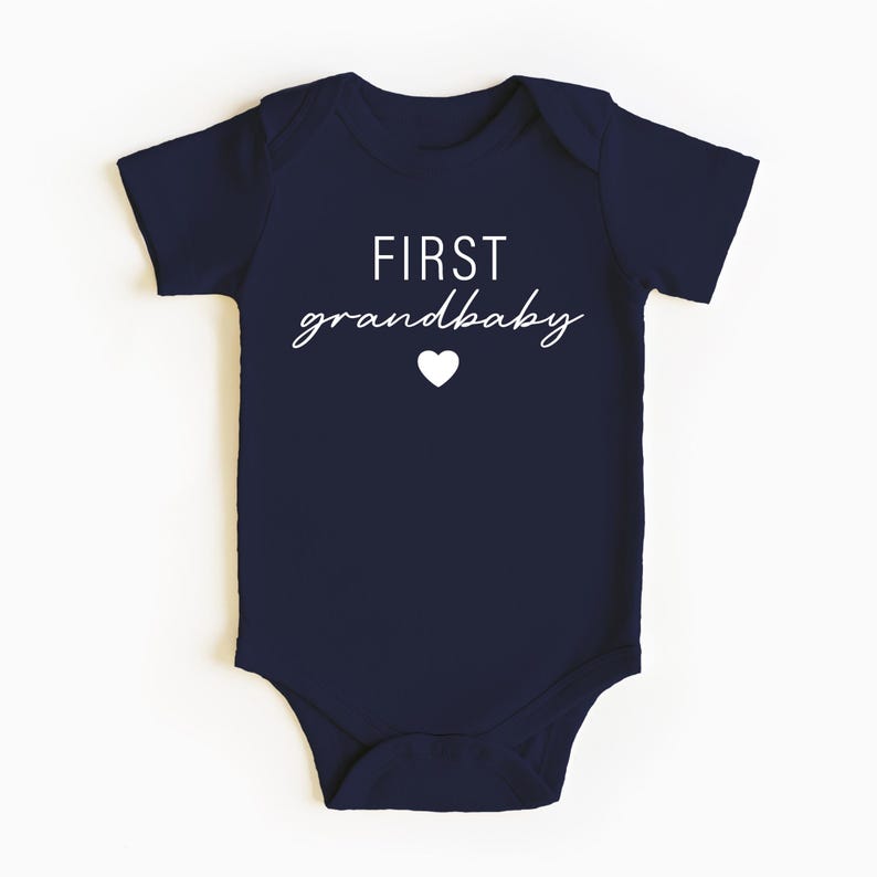 May include: Navy blue baby onesie with white text that reads "FIRST grandbaby" and a small white heart.