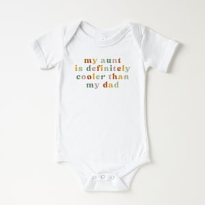 May include: White baby onesie with the text "my aunt is definitely cooler than my dad" in colorful letters.