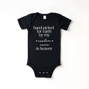 以下が含まれることがあります： 白い文字で「hand picked for earth by my custom name in heaven」と書かれた黒いベビーロンパースです。