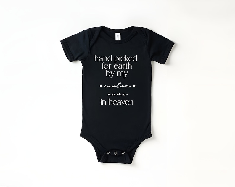 以下が含まれることがあります： 白い文字で「hand picked for earth by my custom name in heaven」と書かれた黒いベビーロンパースです。
