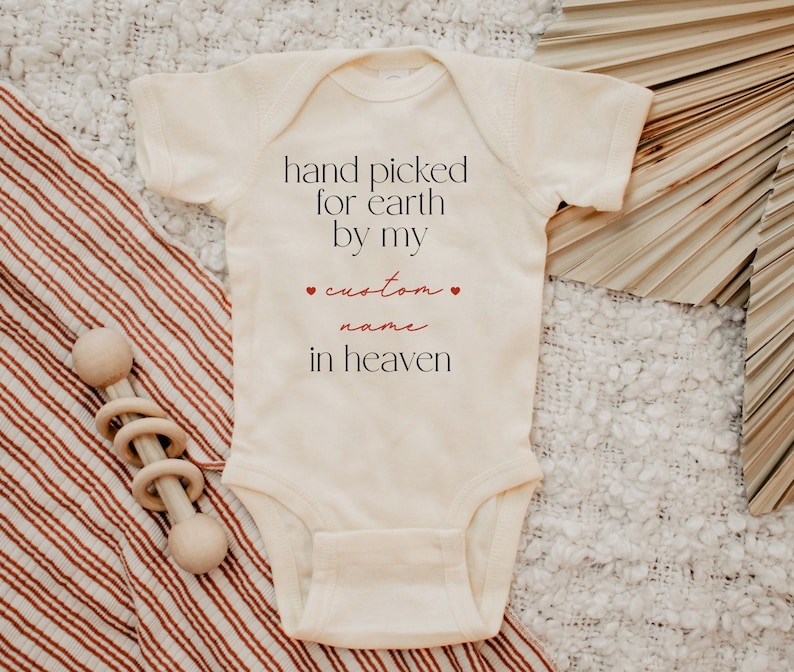 以下が含まれることがあります： 白いベビーロンパースに「hand picked for earth by my custom name in heaven」という赤い文字が書かれています。ロンパースは白と茶色のストライプの毛布の上に置かれています。