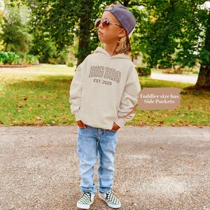 K&ouml;nnte beinhalten: Ein hellbeiger Kapuzenpullover mit dem Text "BIG BRO EST. 2025" in Braun. Ein Kind tr&auml;gt den Pullover mit einer blauen Jeans und schwarz-wei&szlig; karierten Turnschuhen.