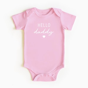May include: Pink baby onesie with white text that reads "HELLO daddy" and a small white heart.