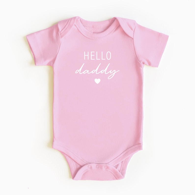 May include: Pink baby onesie with white text that reads "HELLO daddy" and a small white heart.