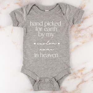 以下が含まれることがあります： 「hand picked for earth by my custom name in heaven」というテキストが書かれたグレーのベビーロンパース。