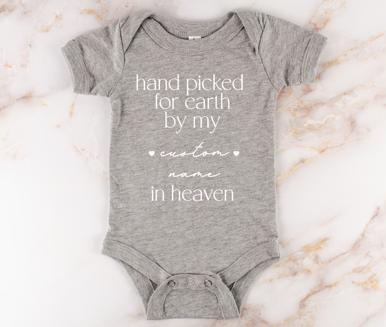 以下が含まれることがあります： 「hand picked for earth by my custom name in heaven」というテキストが書かれたグレーのベビーロンパース。