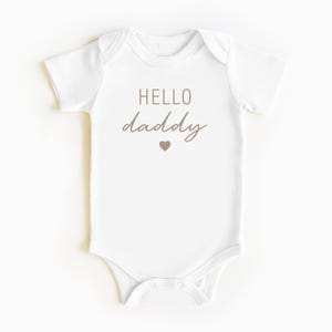 May include: White baby onesie with brown text that says "HELLO daddy" with a small brown heart below the text.