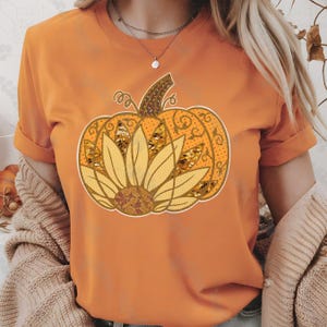 May include: Orange t-shirt featuring a pumpkin design with a sunflower in the center. The pumpkin is decorated with swirls and dots, and the sunflower has yellow petals. The shirt has short sleeves and a round neckline.