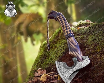 May include: A handcrafted axe with a wooden handle featuring a woven leather grip. The axe head is silver-colored with intricate engraved designs. A leather cord is attached to the handle. The axe is displayed on a moss-covered surface.