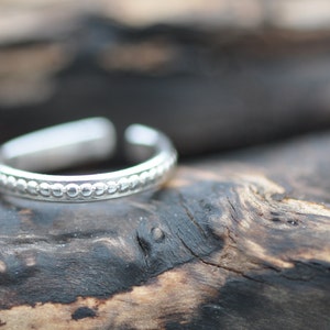May include: A silver toe ring with a beaded design. The ring is on a piece of weathered wood.