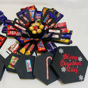 May include: A black hexagonal explosion box filled with various chocolate bars and sweets, including Kinder, Twix, Dairy Milk, and Cadbury. The box has a lid with a red inscription that reads "Merry Christmas Riley".